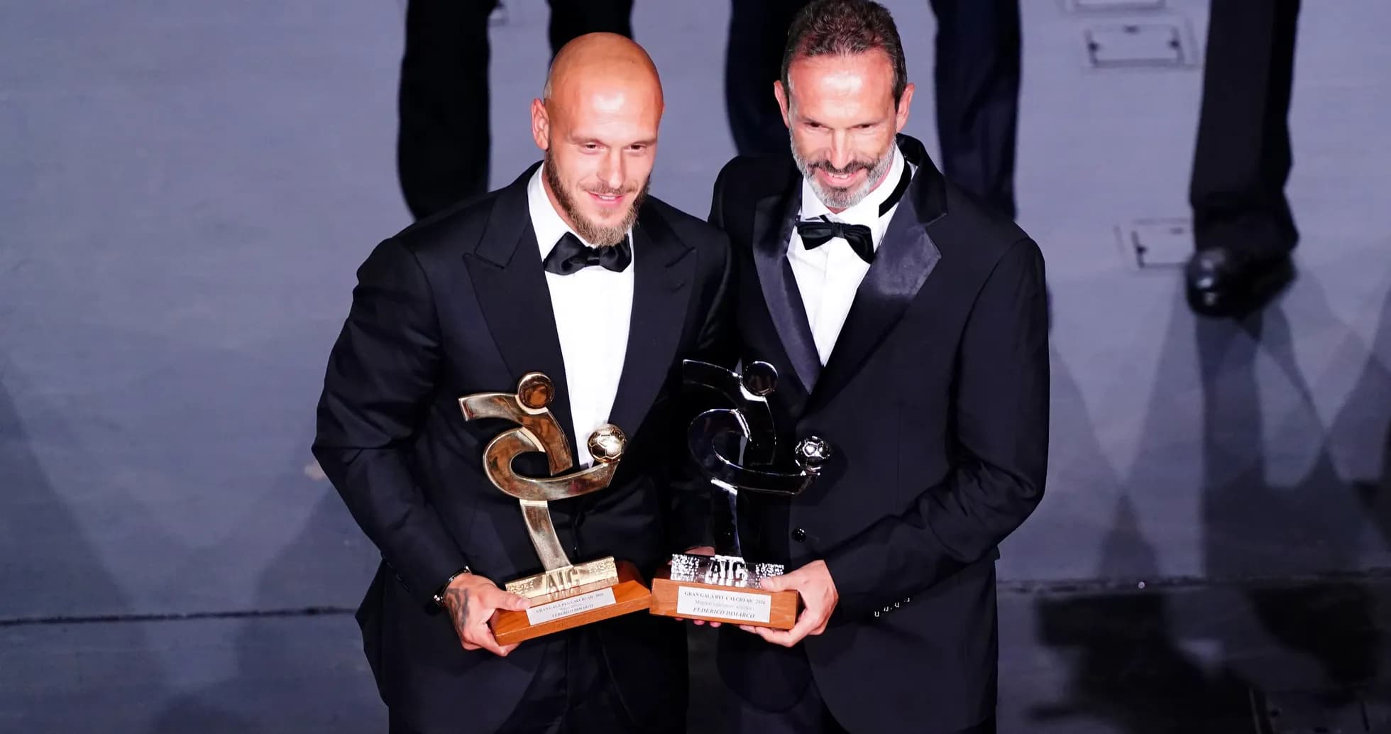 Two men in tuxedos holding awards on stage Two men in tuxedos holding awards on stage